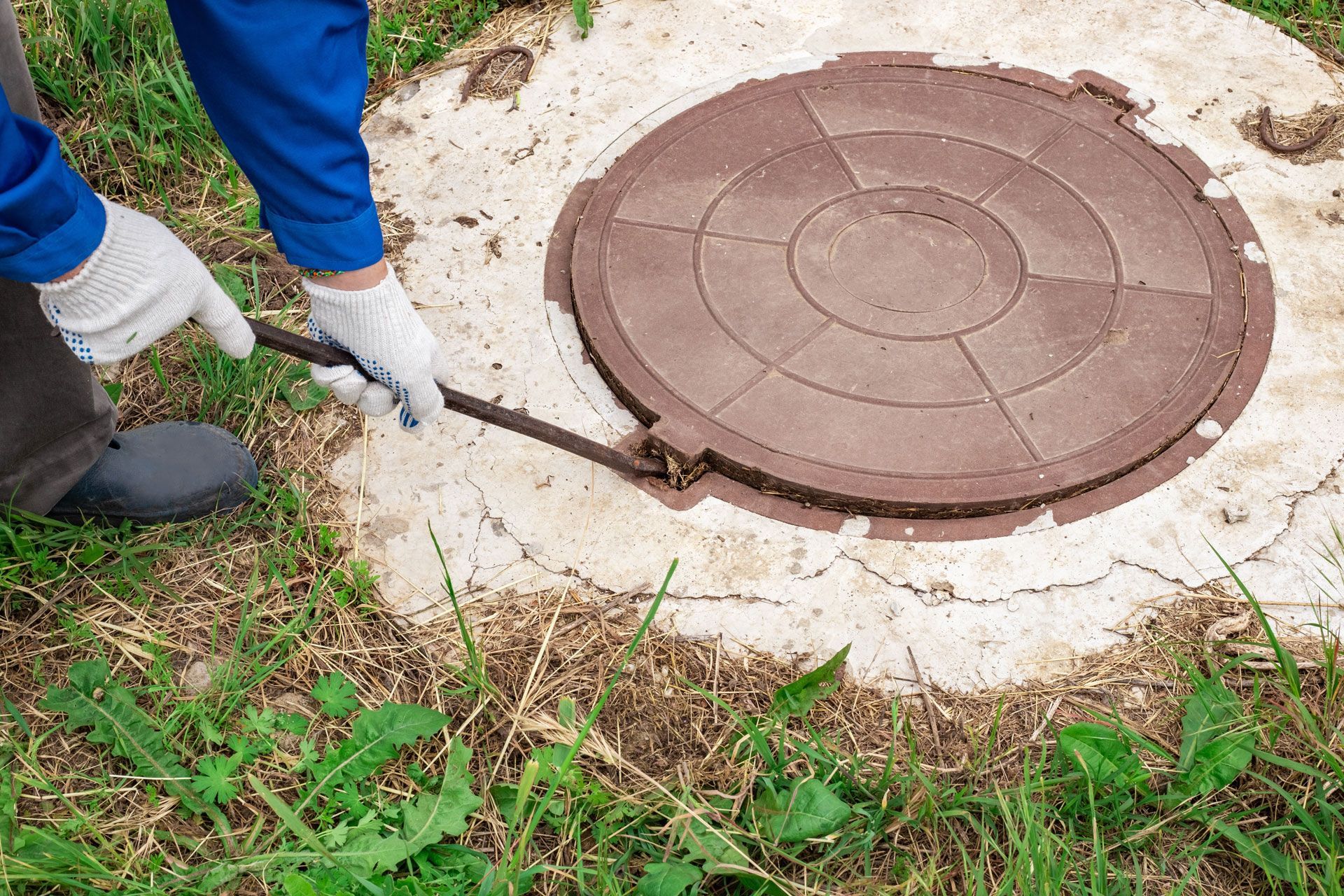 Person in gloves using a tool to lift a brown manhole cover set in concrete, surrounded by grass.