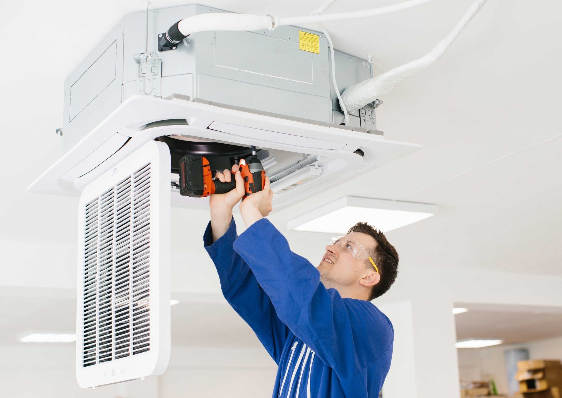 Man in blue uniform using a drill to work on ceiling-mounted HVAC unit. White room setting.