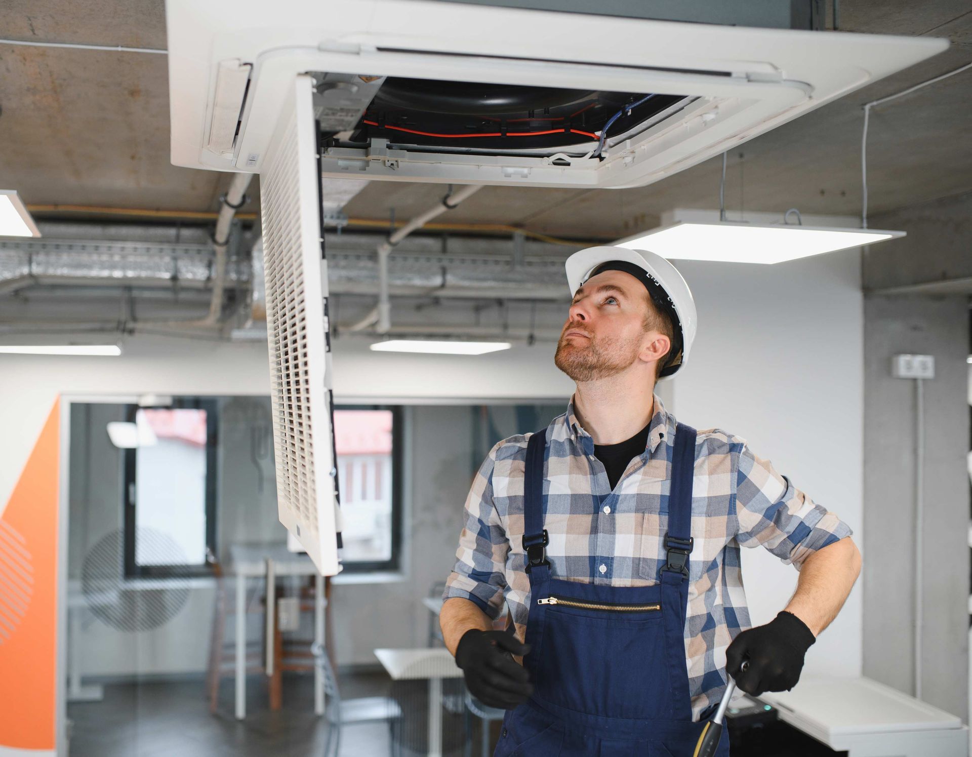 HVAC technician in overalls and hard hat inspecting a ceiling air conditioner unit. Indoors.