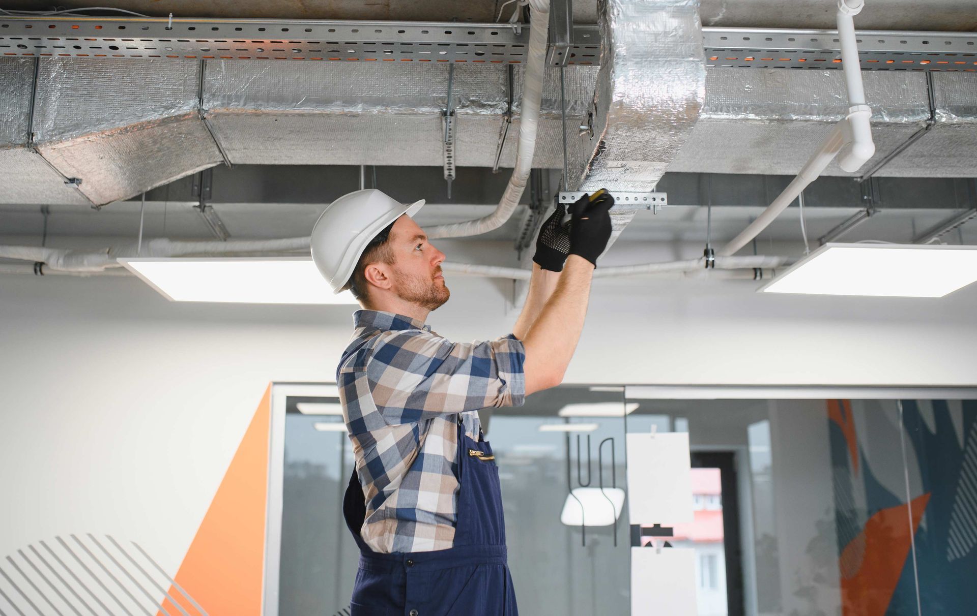 Man in hard hat and work clothes installing ductwork in an office ceiling