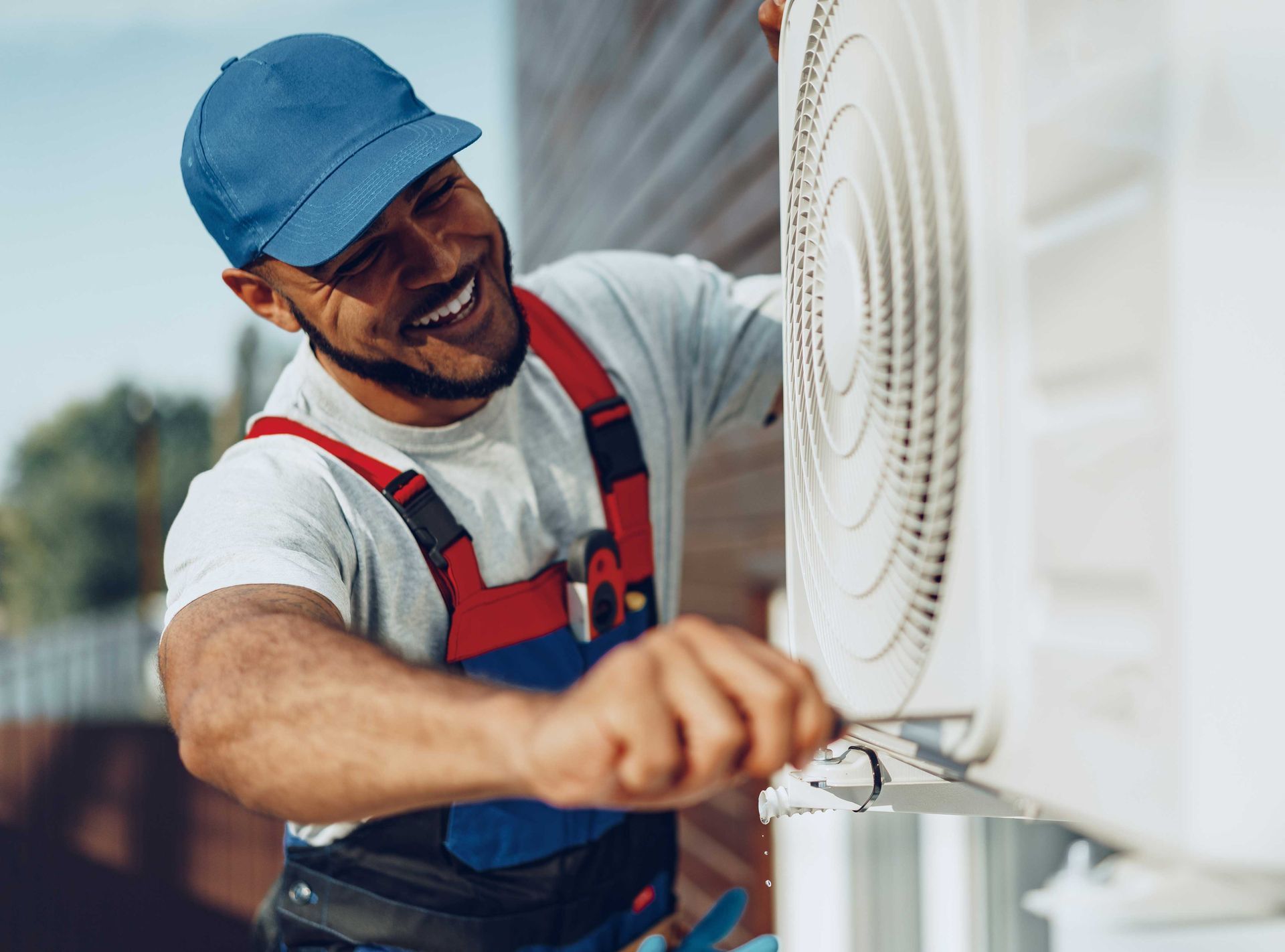 HVAC technician in a blue cap smiles while working on an air conditioner unit outdoors.