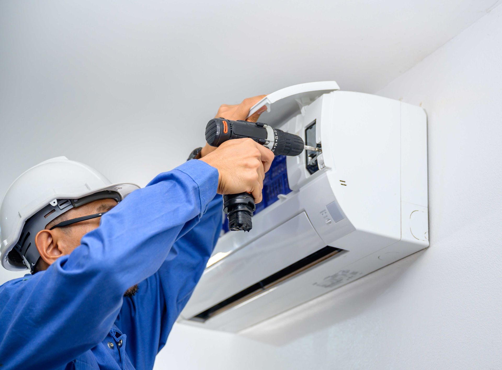 HVAC technician installing an air conditioner unit on a white wall using a power drill.