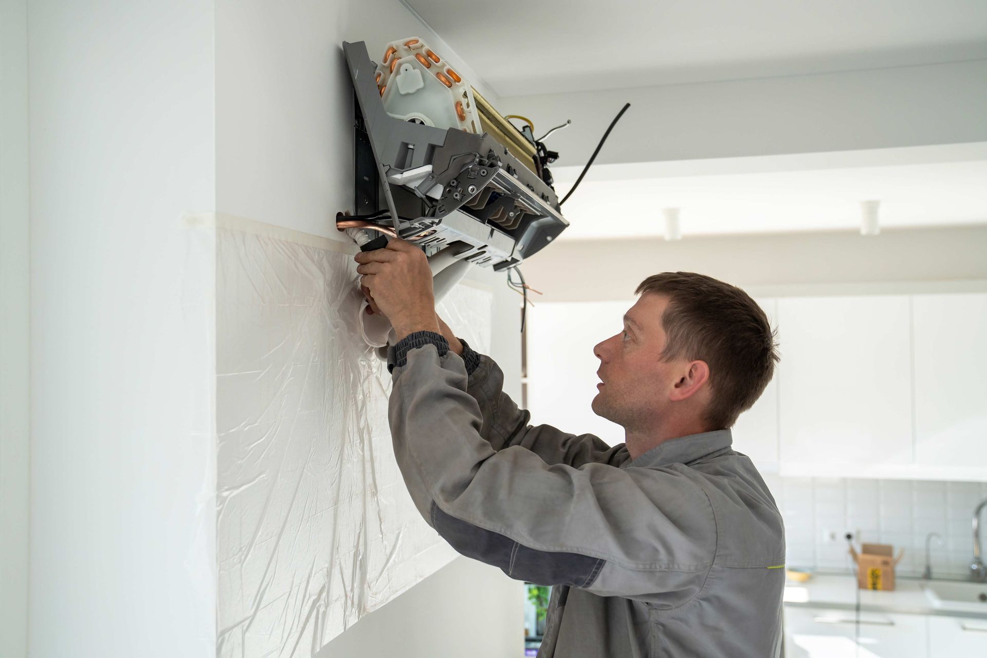 Man in gray shirt installing equipment on a white wall in a modern kitchen.