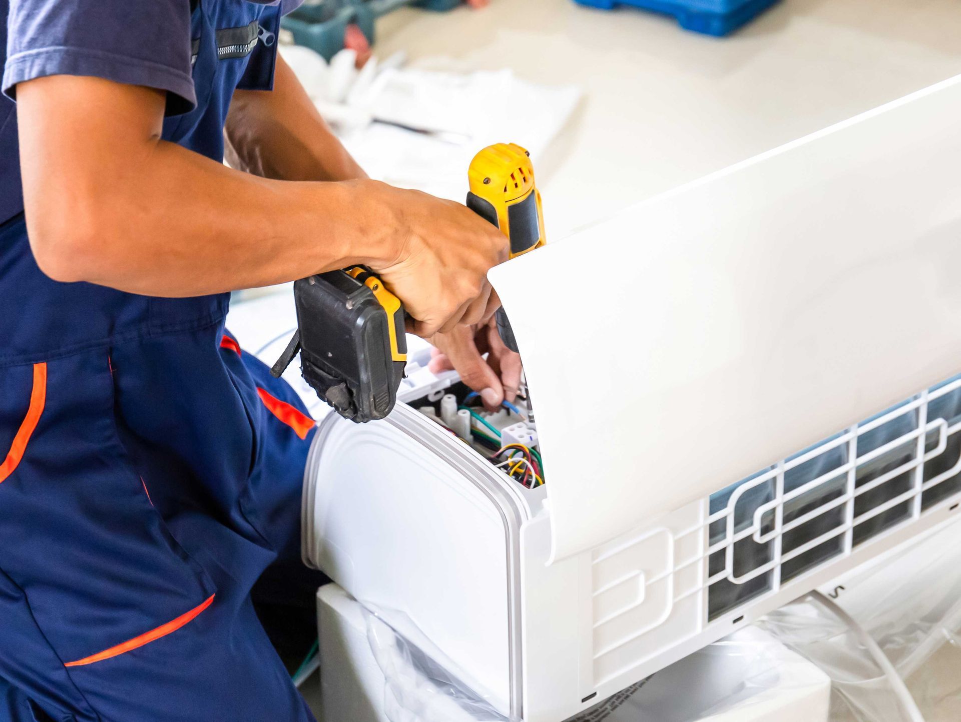 Technician in blue overalls uses a drill to repair a white air conditioning unit.