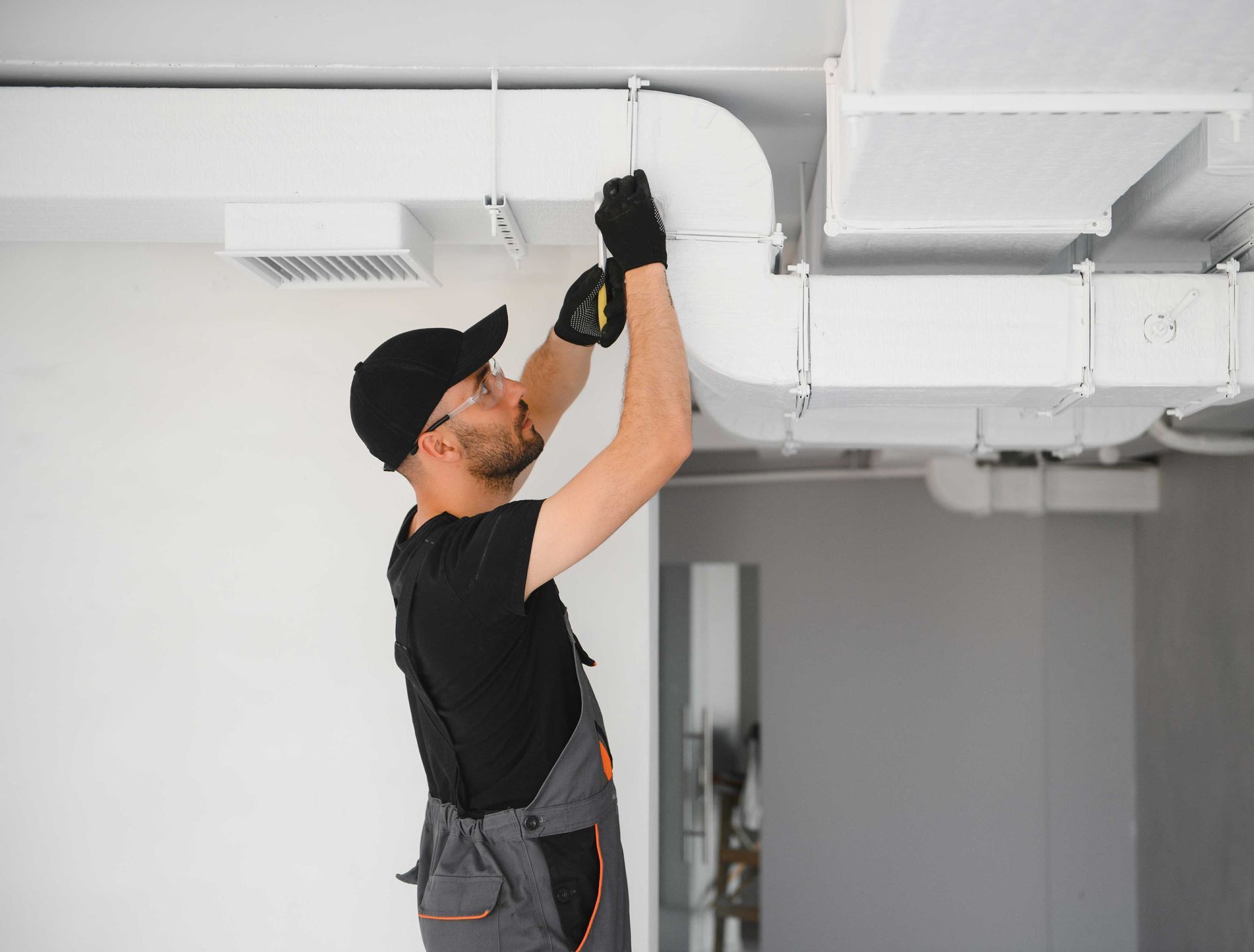 HVAC technician in work clothes, installing ductwork on a white ceiling.
