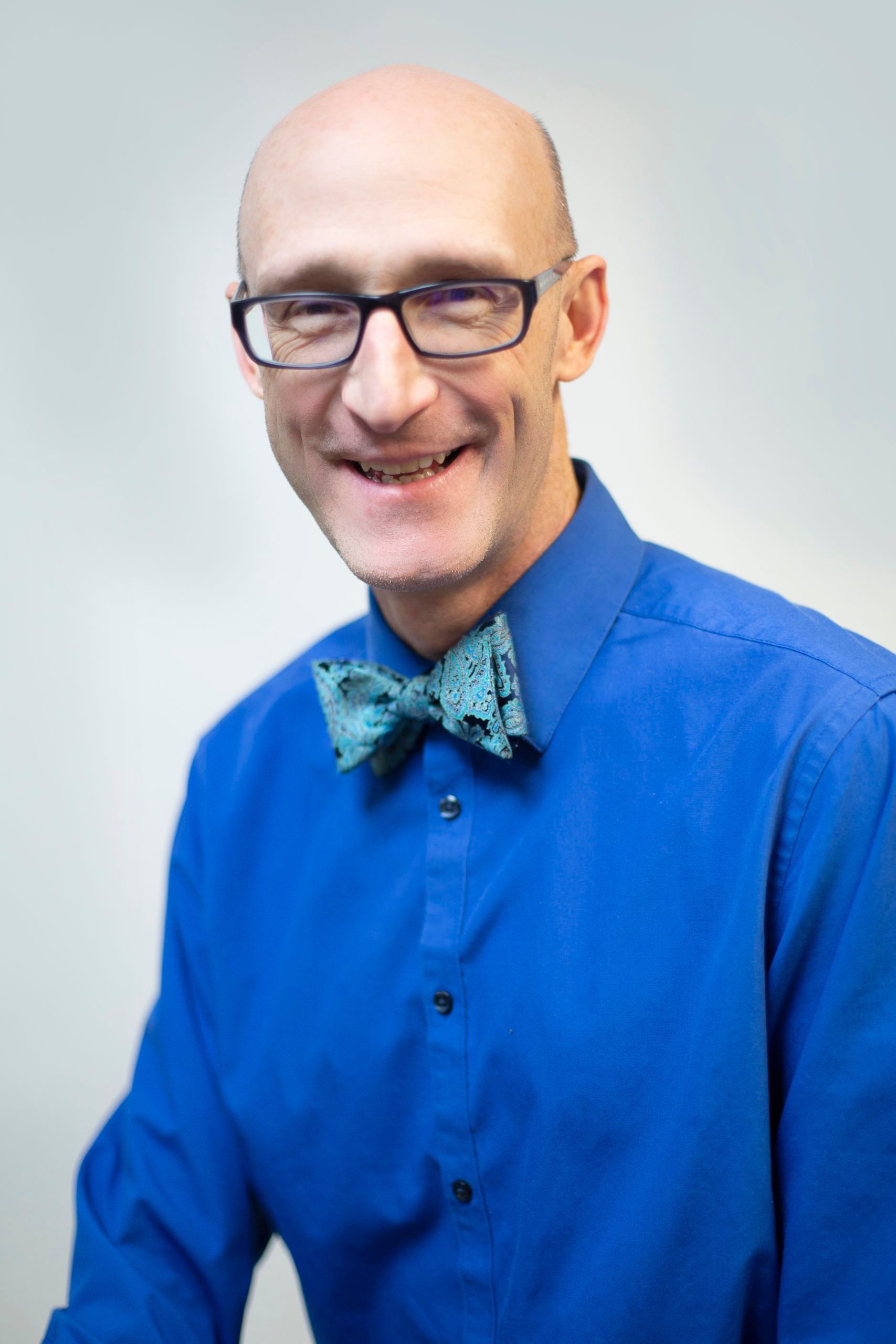 A bald man wearing glasses and a blue shirt with a green bow tie.