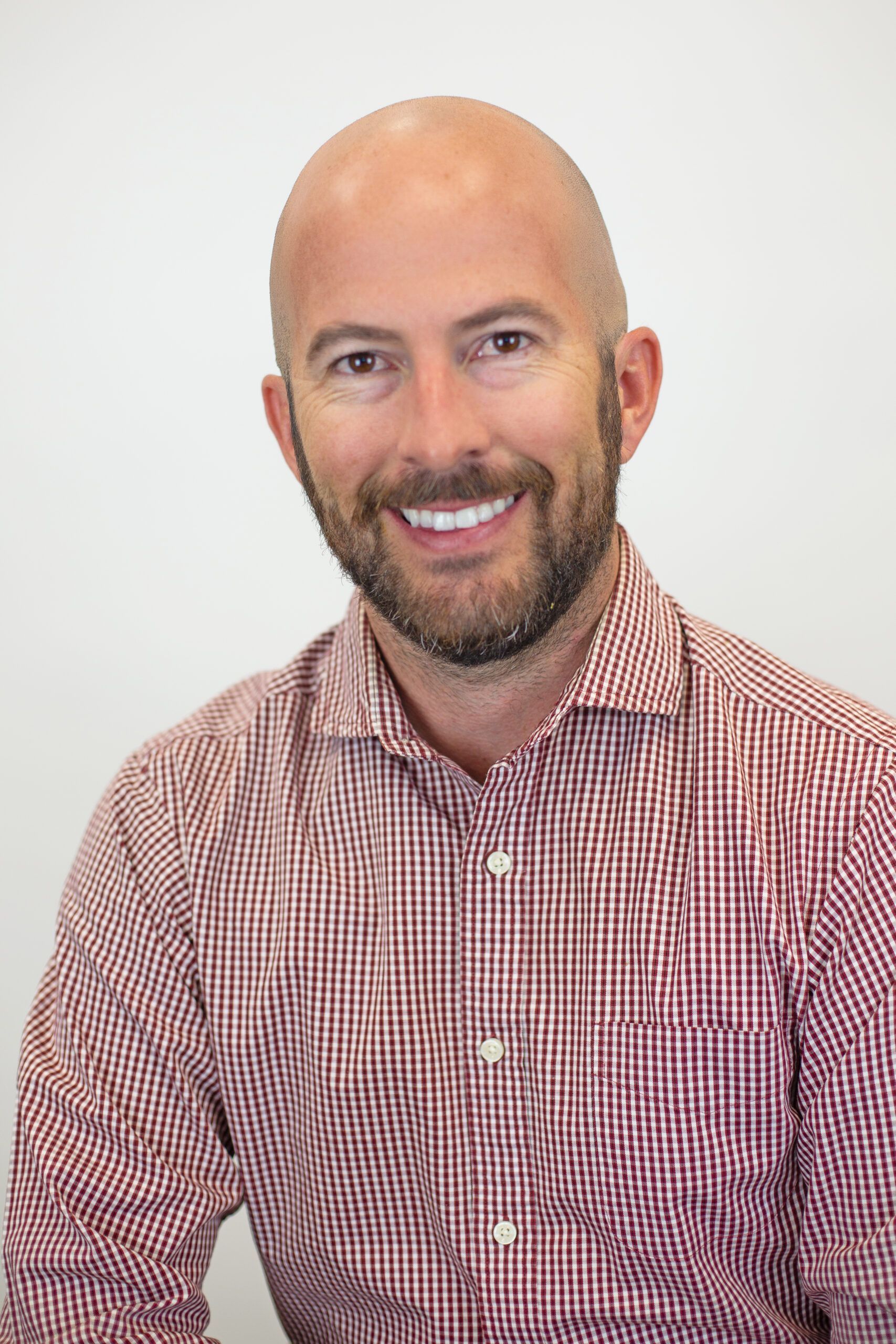 A bald man with a beard is wearing a red and white plaid shirt and smiling.