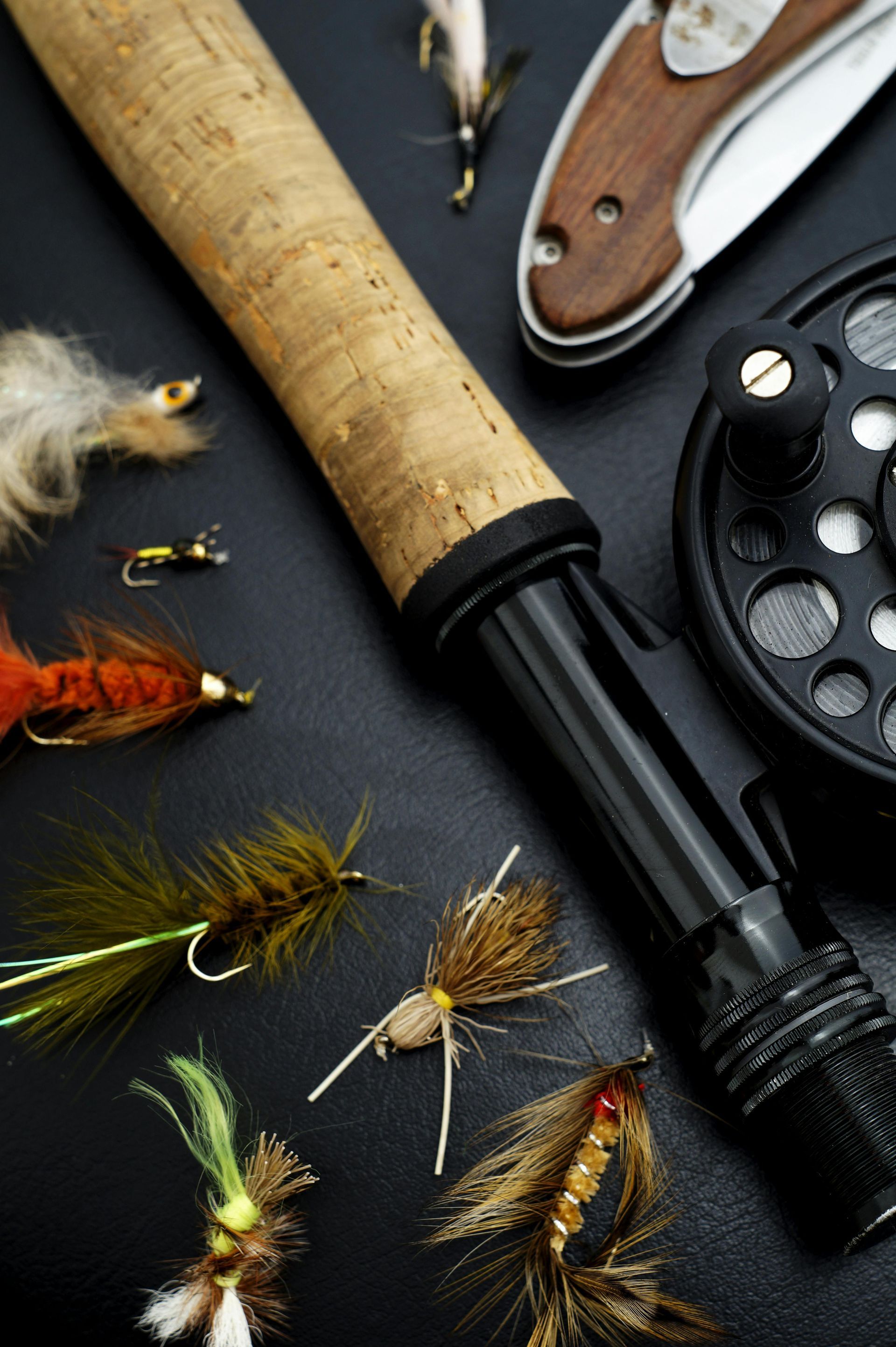 Fly fishing rod and reel with various colorful fishing flies, and a pocket knife on a black surface.