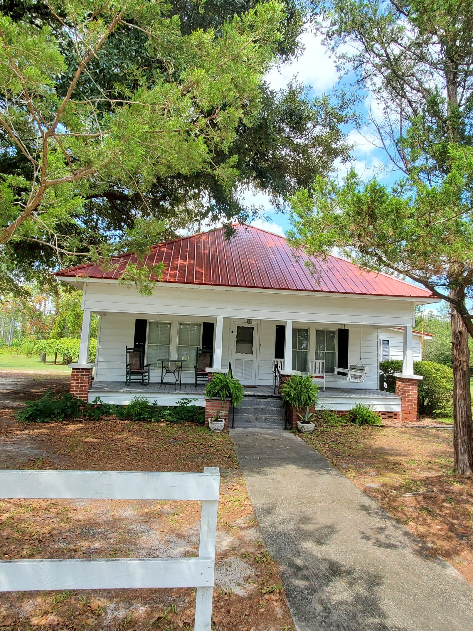 White house with a red roof, a porch, and a white fence in a yard surrounded by trees.