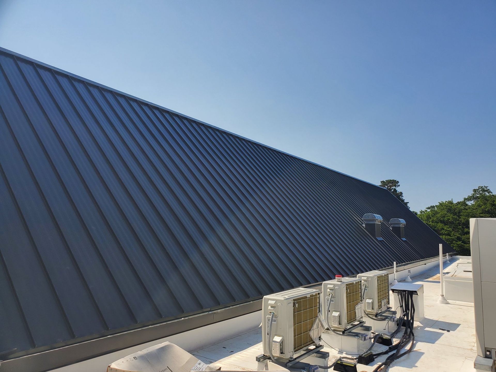 Dark metal roof on a building, with air conditioning units below and a clear blue sky.