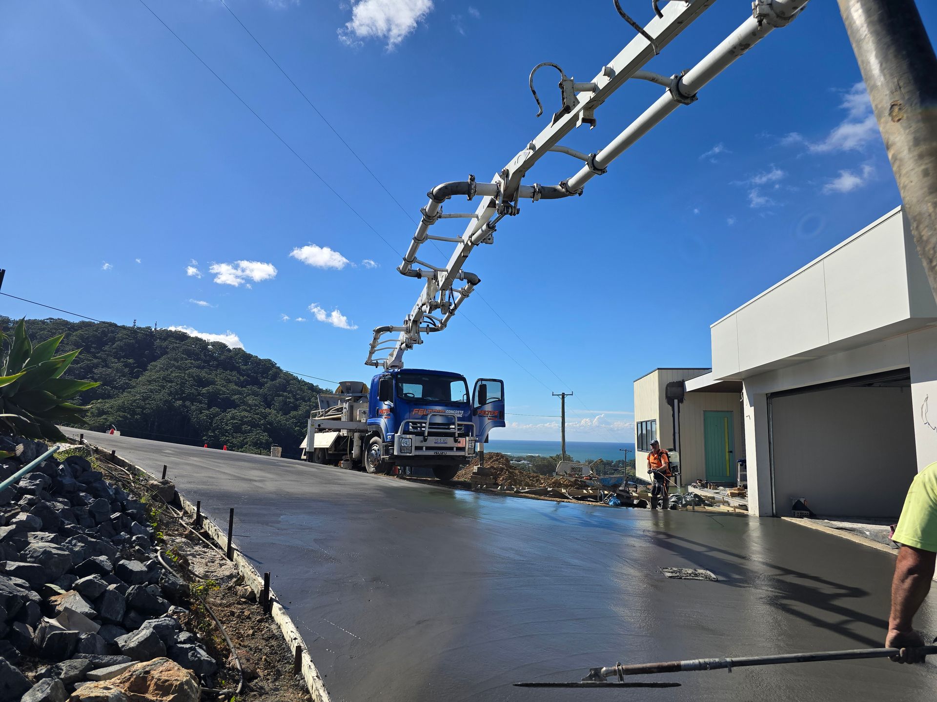 A concrete pump truck pouring concrete on a driveway with an ocean view on a sunny day.