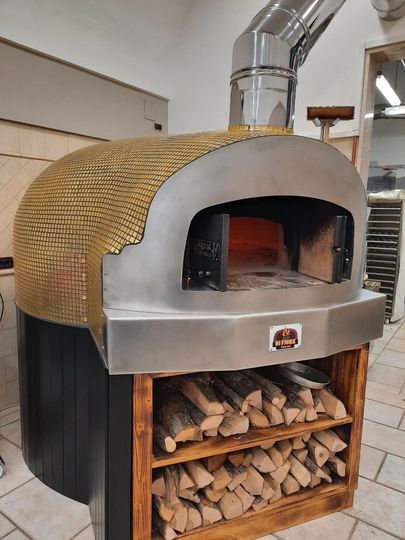 Forno per pizza a legna con focolare a vista e legna da ardere accatastata al di sotto.