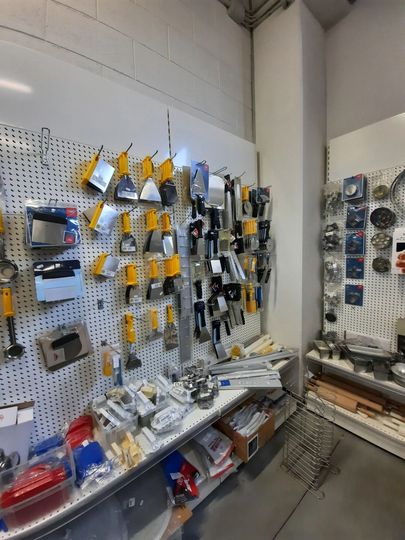 Parete forata con attrezzi e componenti vari in un corridoio di un'officina, con scaffali e materiali sottostanti.