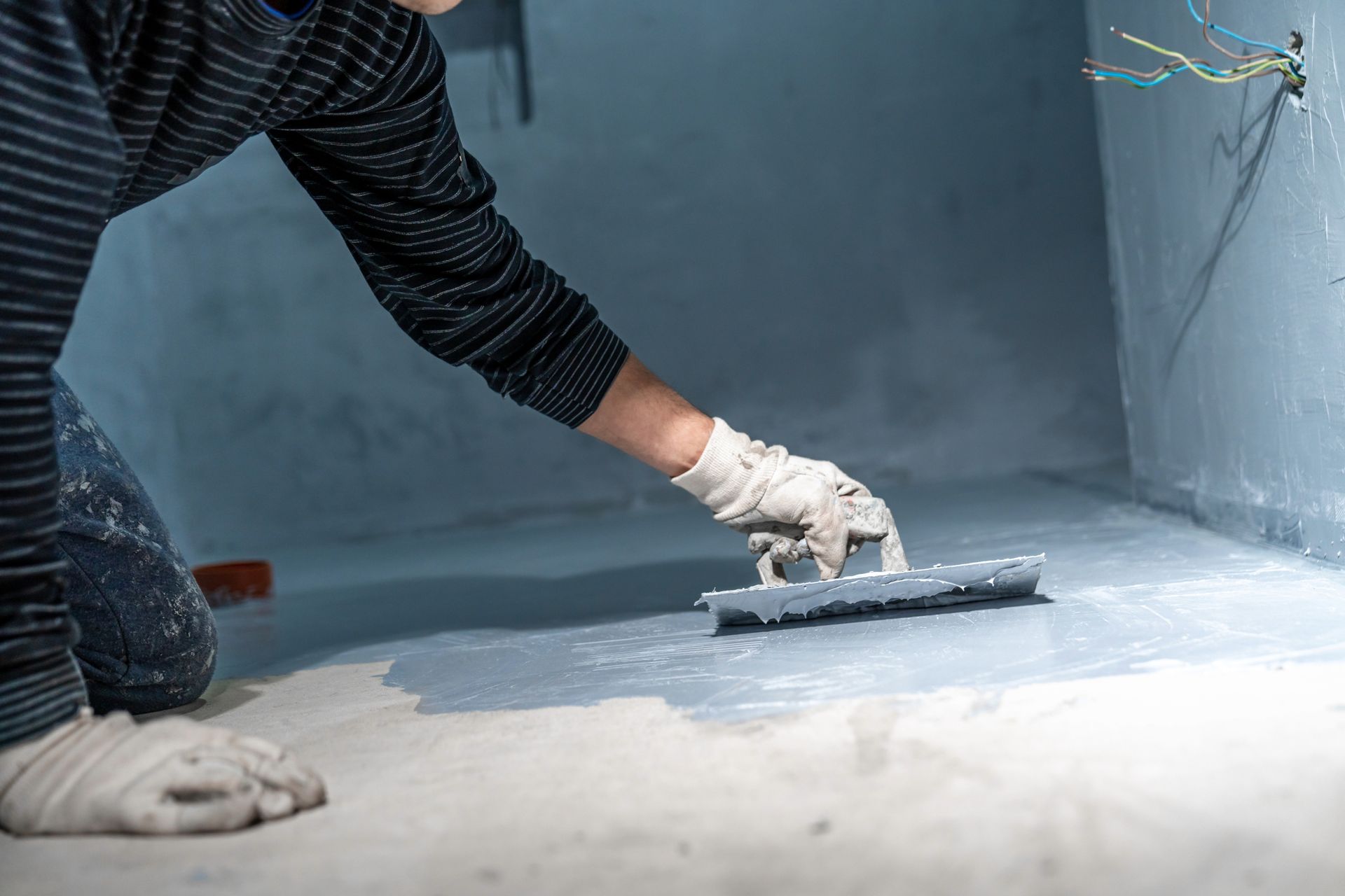 A person wearing gloves uses a trowel to apply gray waterproof coating to a floor in a room under construction.