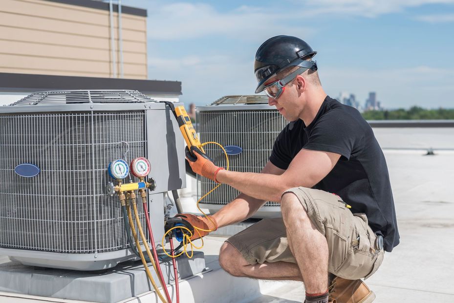 Technician kneeling beside rooftop HVAC unit, checking gauges and wiring with a tool.