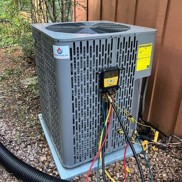 An air conditioning unit with gauges attached, outside a building, surrounded by foliage.