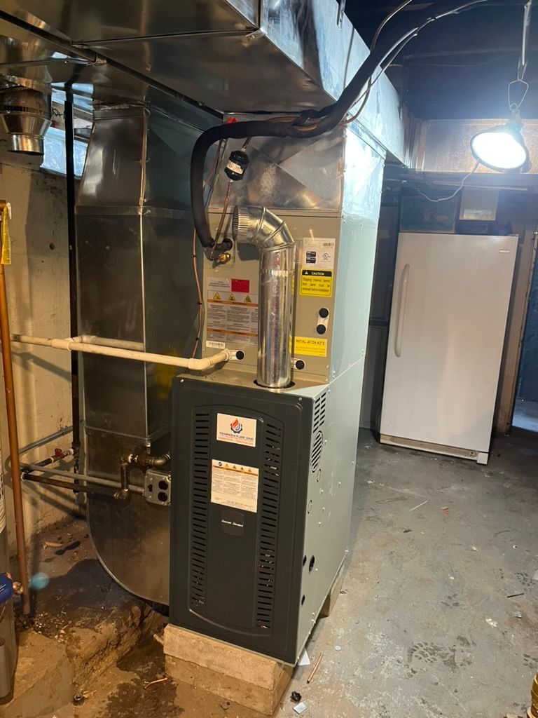 Furnace with ductwork in a basement. Grey metal unit with labels. White refrigerator in the background.