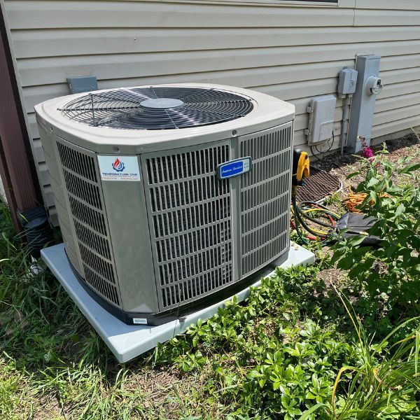 Gray air conditioning unit outside a light-colored building, on a concrete pad, surrounded by grass and plants.