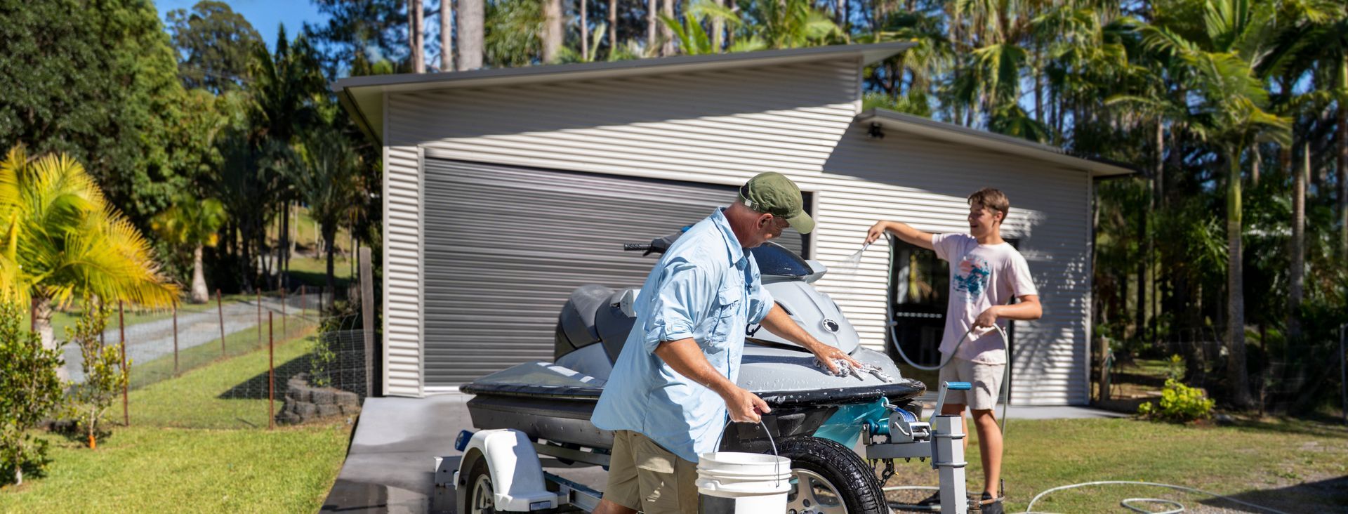 A man and a boy washing a jet ski on a trailer in front of a garage.