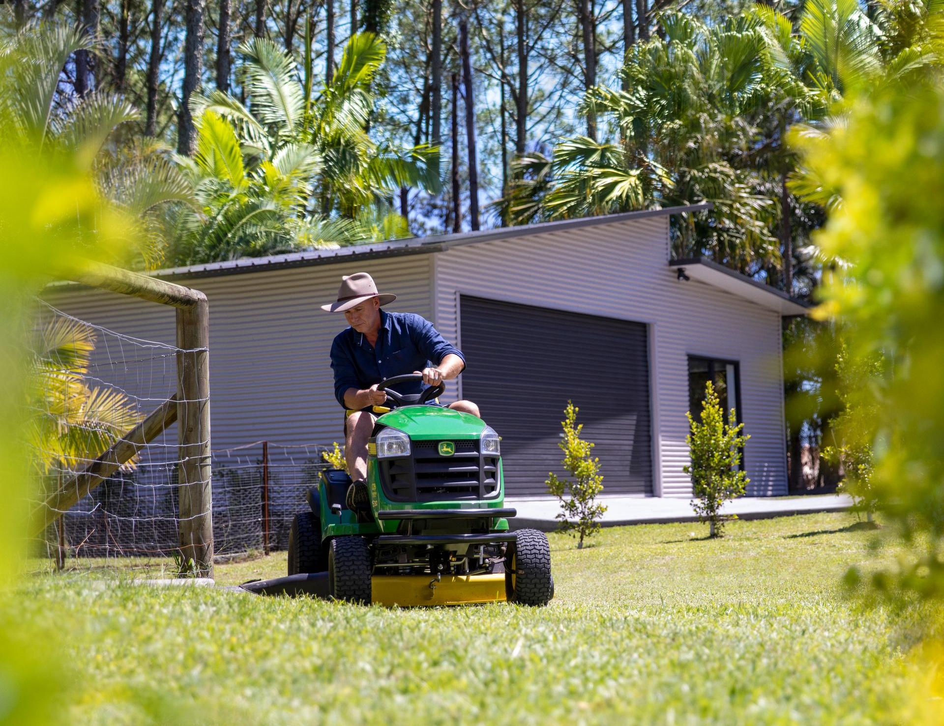 Man mowing the lawn on a John Deere tractor in a sunny yard with a shed in the background.