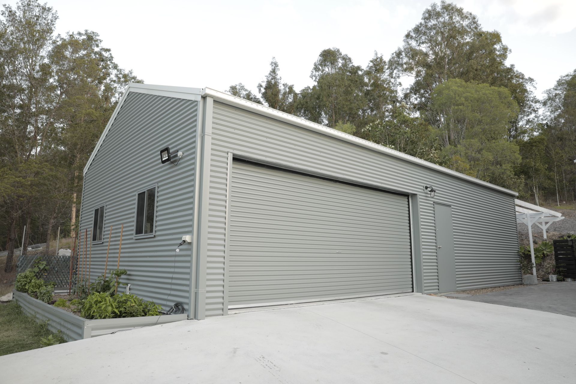 Silver metal garage with large door and concrete driveway, surrounded by trees.