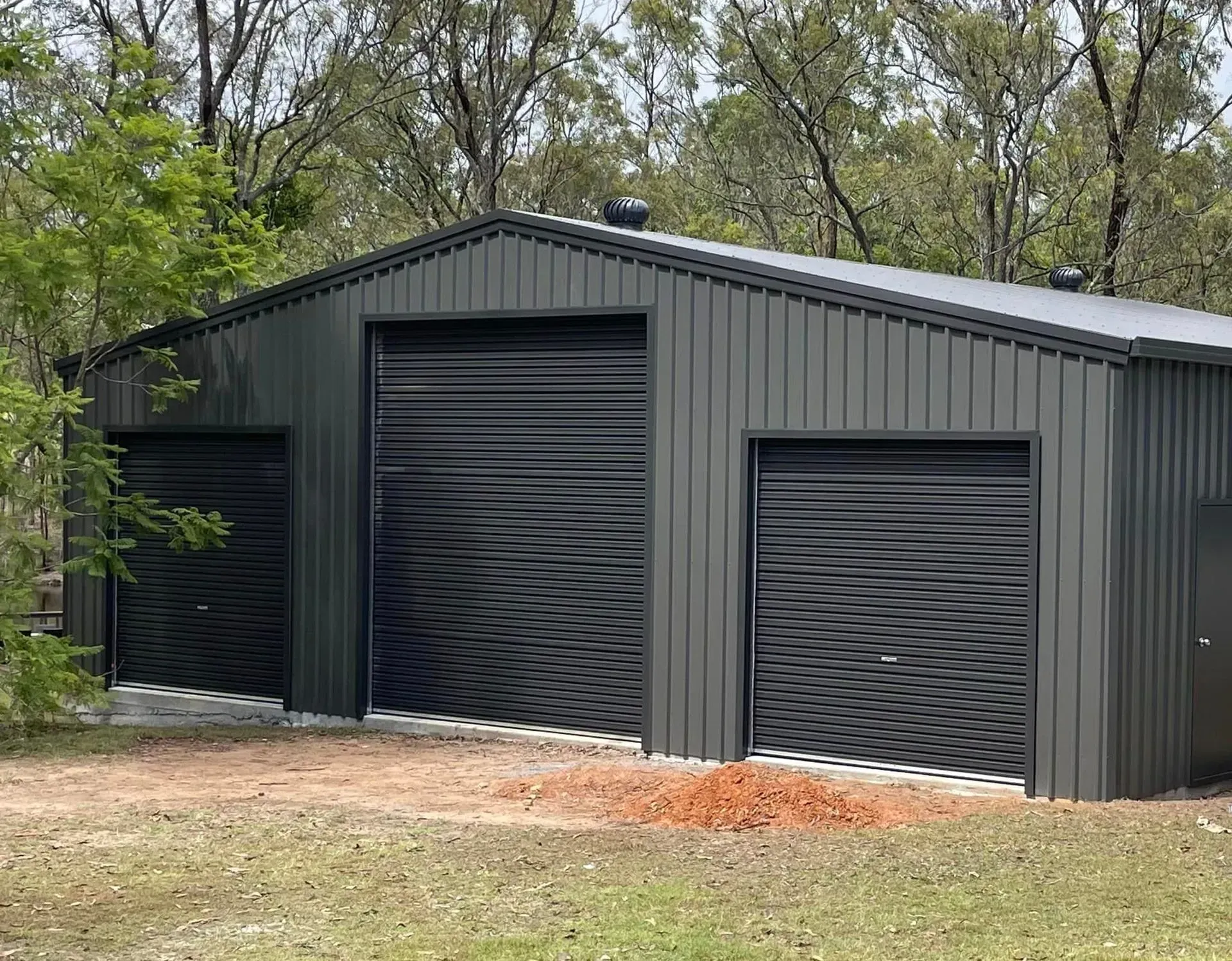 Dark Gray Metal Shed with Three Roll-Up Doors, Set in A Grassy Area — Fair Dinkum Builds Port Stephens in Bobs Farm, NSW