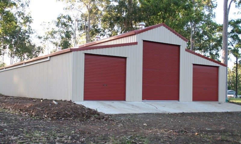 Tan metal shed with three red garage doors. Located outdoors, surrounded by trees.