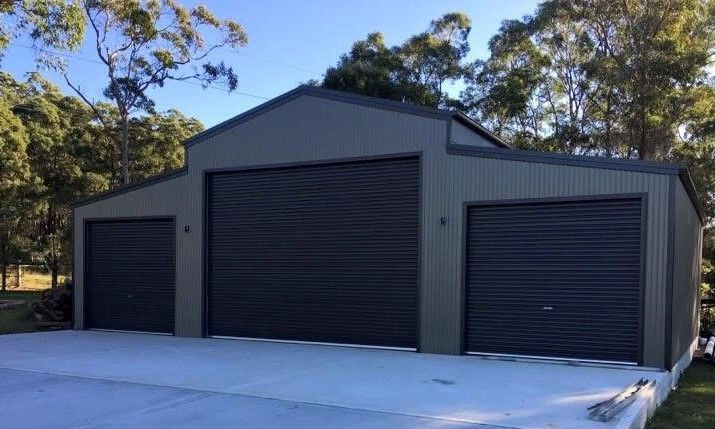 Grey metal shed with three roller doors, on a concrete slab, in a wooded area.