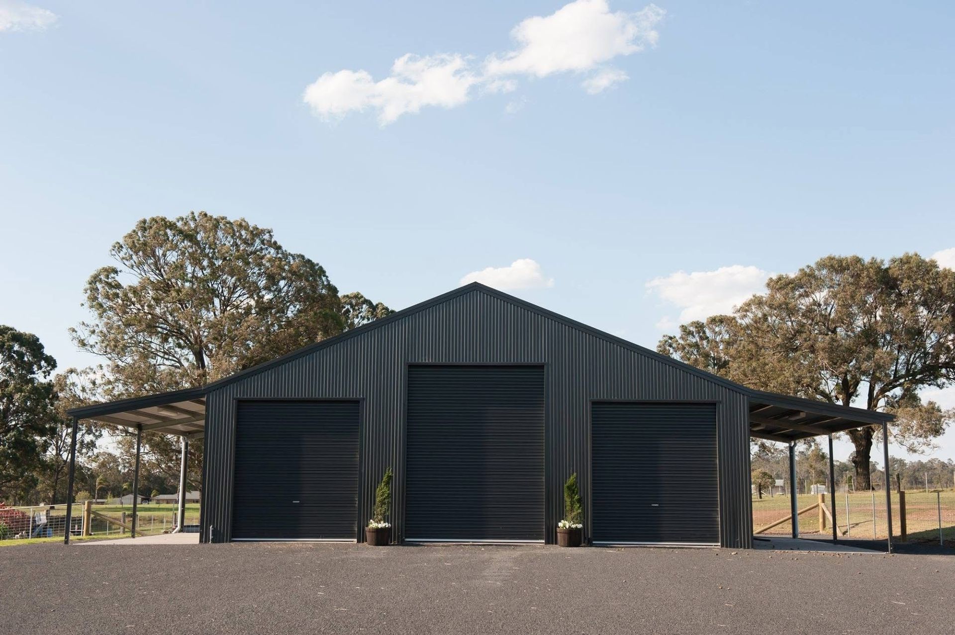Black metal garage with three bays, under a clear sky, surrounded by trees.