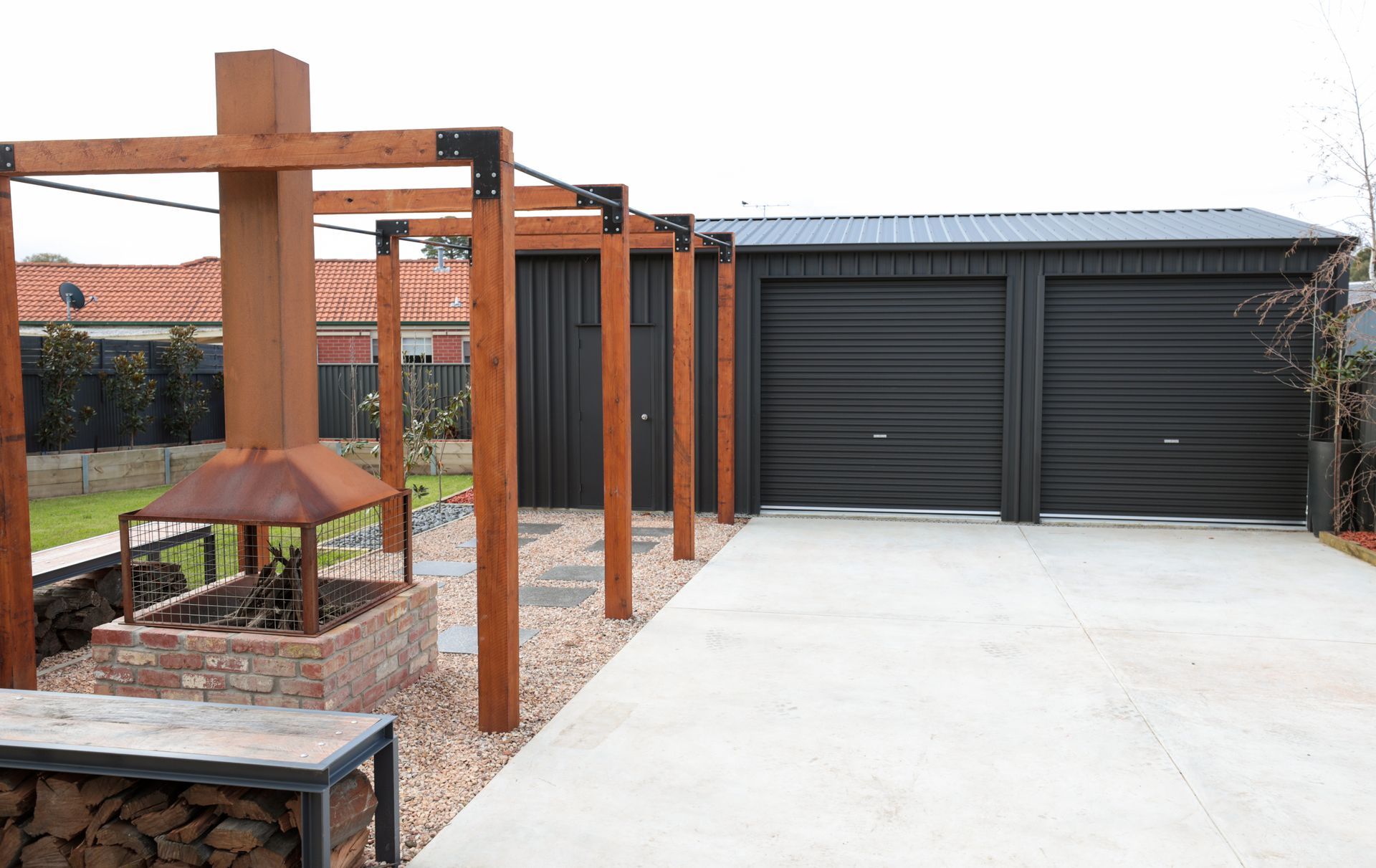 Exterior view: black garage with two doors, wooden pergola, fire pit, and gravel pathway.