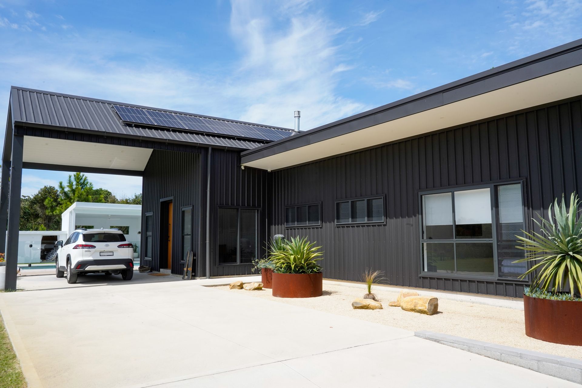 Dark gray modern building with solar panels, a car under a carport, and landscaping.