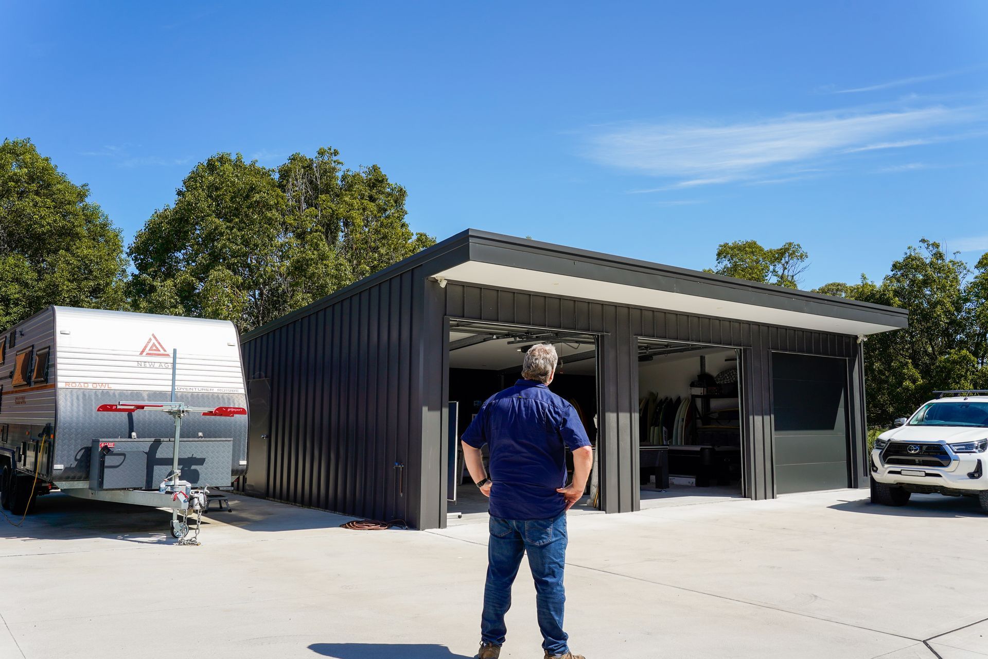 Man standing in front of dark gray shed with open doors; caravan and white truck nearby, under a blue sky.