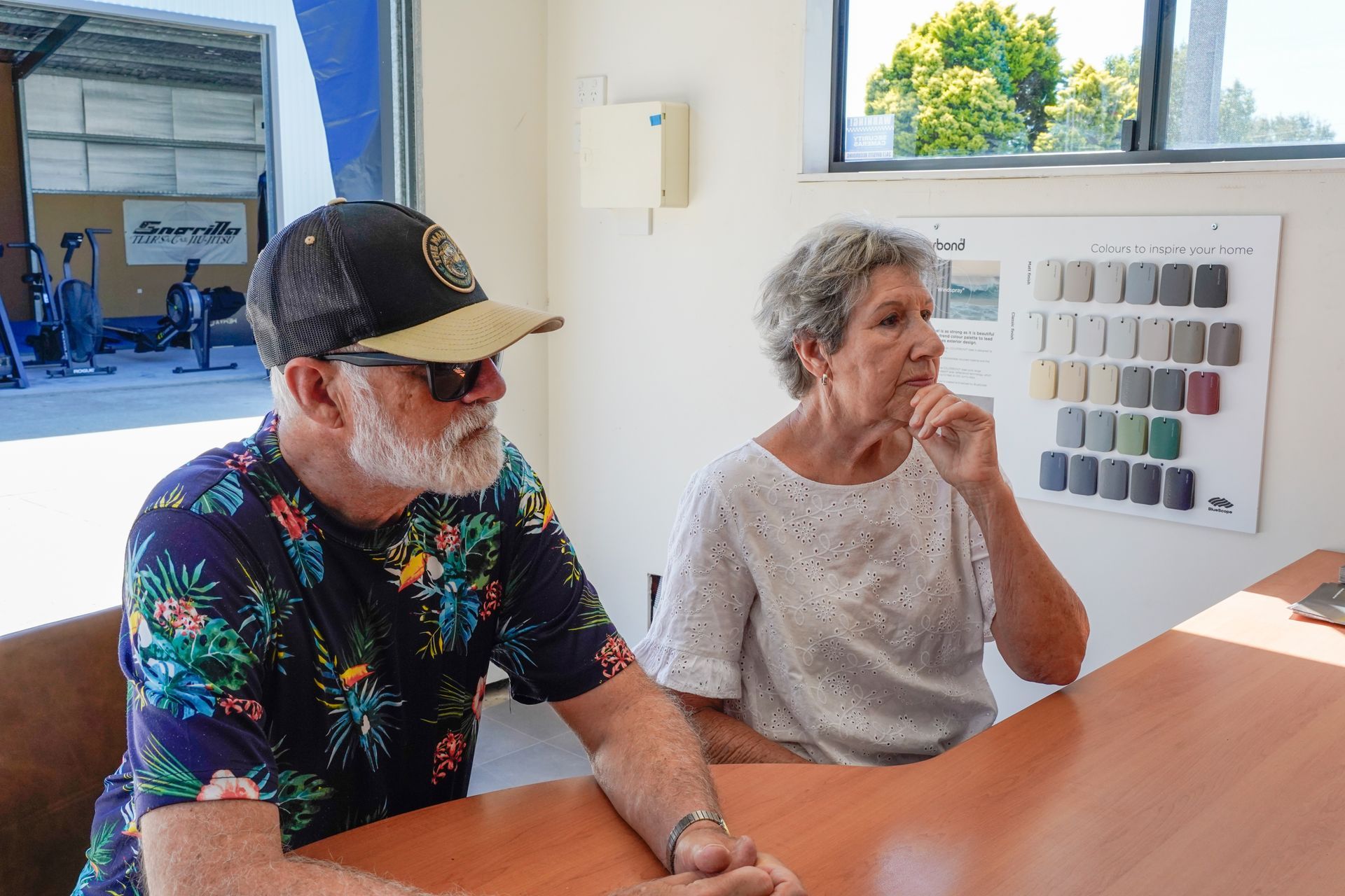 A man and woman sit at a table looking at something out of frame; a color sample chart is visible behind them.