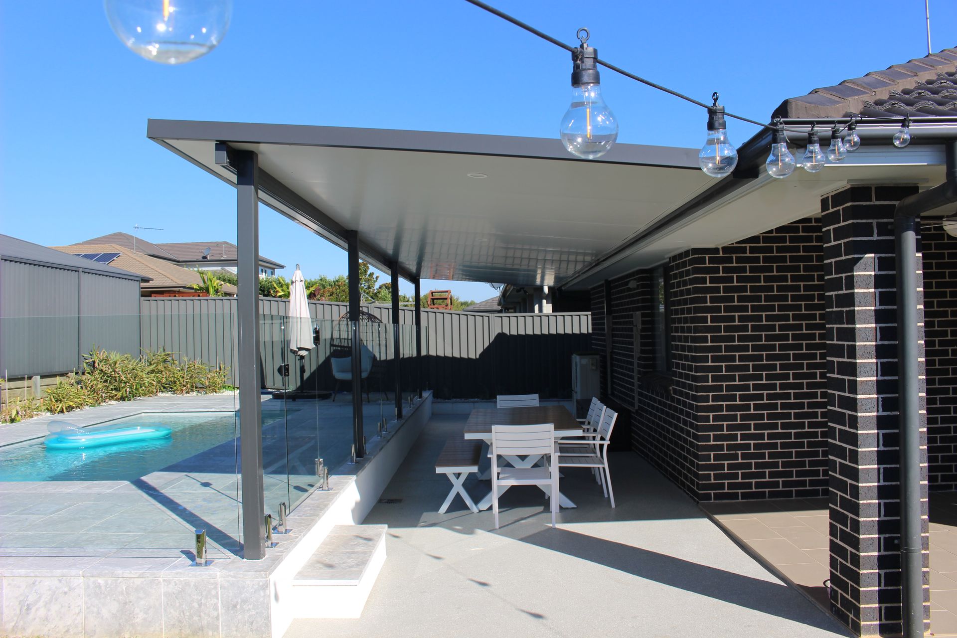 Patio with table, chairs, and pool. Black awning over a dining area. String lights hang above.