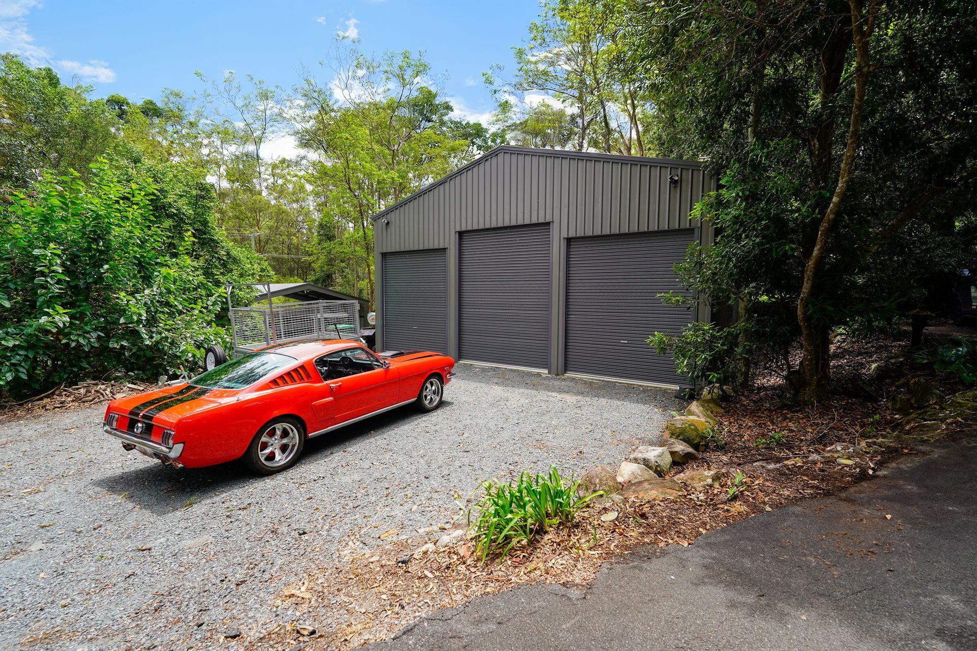 Red classic car parked in front of a gray metal garage. Gravel driveway, surrounded by trees.