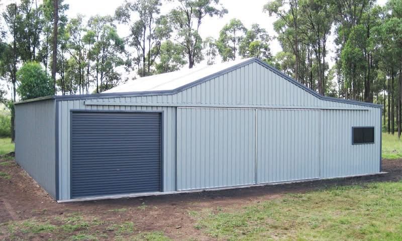 Gray metal shed with a roller door and sliding door, set in a grassy area with trees in the background.