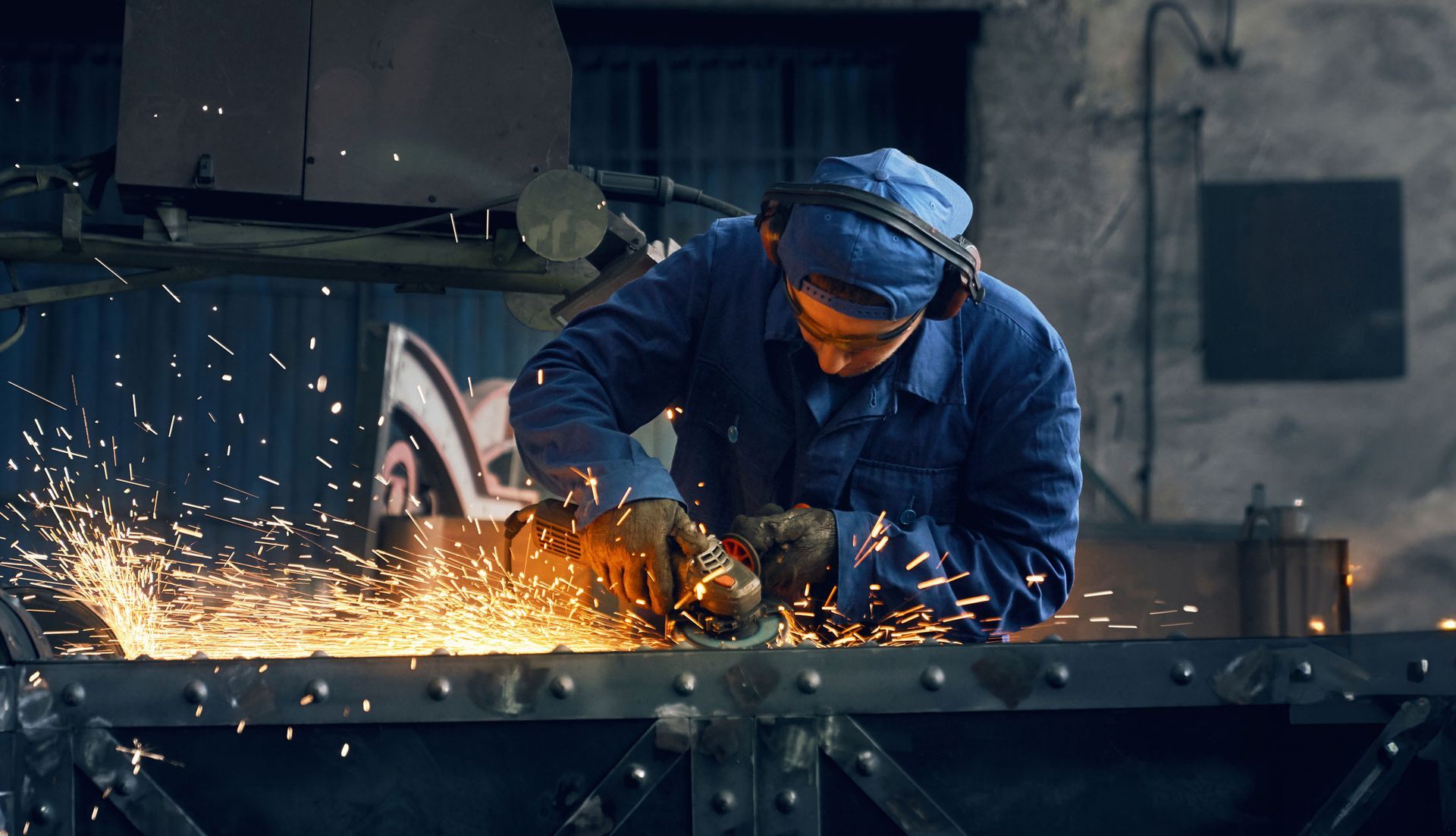 Person in blue workwear grinding metal, sparks flying in an industrial setting.