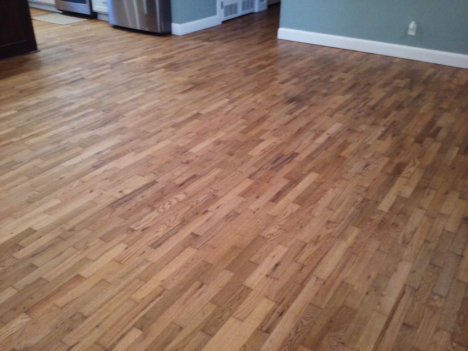 A wooden floor in a living room with a stainless steel refrigerator in the background.
