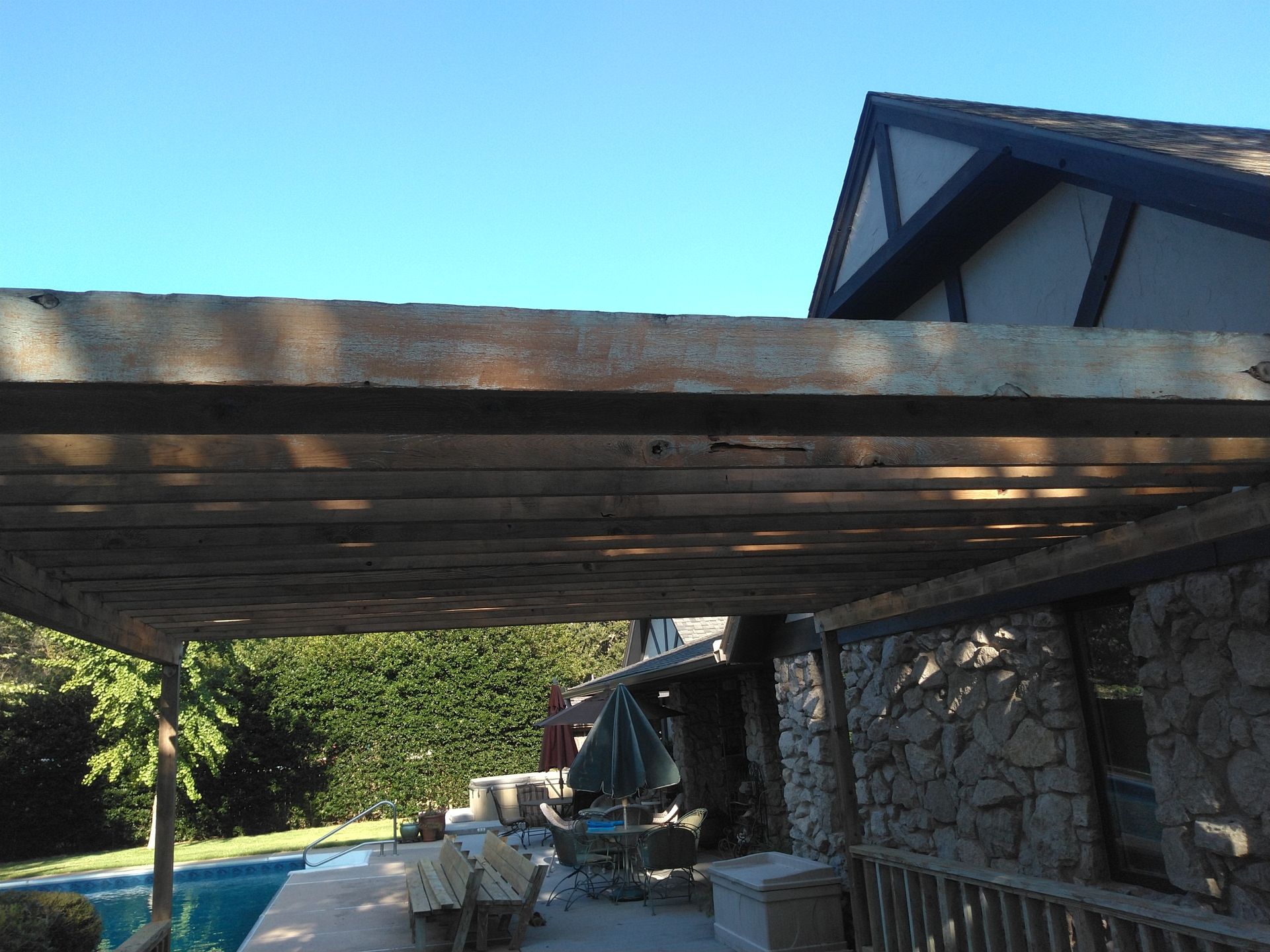 A wooden pergola over a patio with a swimming pool in the background