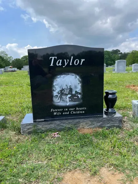 Black memorial headstone engraved “Taylor” with a floral arch, in a grassy cemetery under a cloudy sky