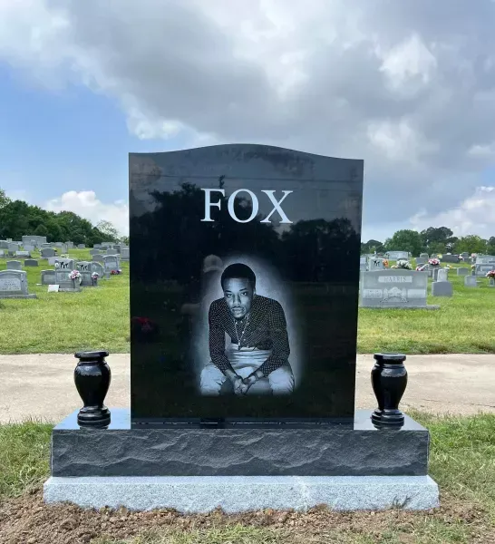 Black gravestone engraved “FOX” with a portrait, set in a cemetery with rows of headstones and cloudy sky
