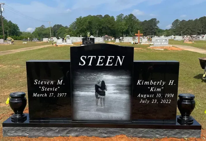 Black STEEN gravestone with portrait photo in a cemetery, flanked by name plaques and urns.