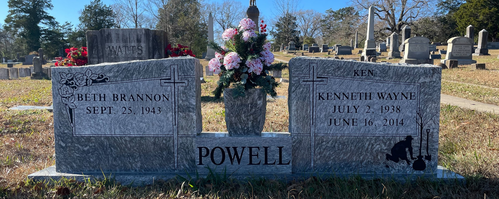 Two gray memorial stones with flowers in a cemetery, marked “POWELL,” under a bright blue sky