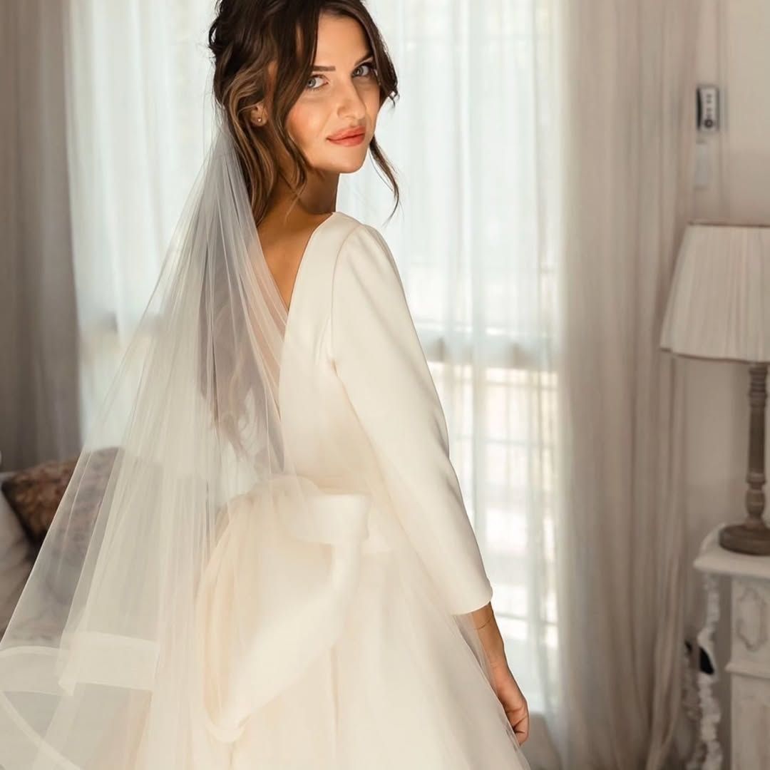 Donna in abito da sposa, che guarda oltre la spalla. In un interno, vicino a una finestra. Abito beige con velo.