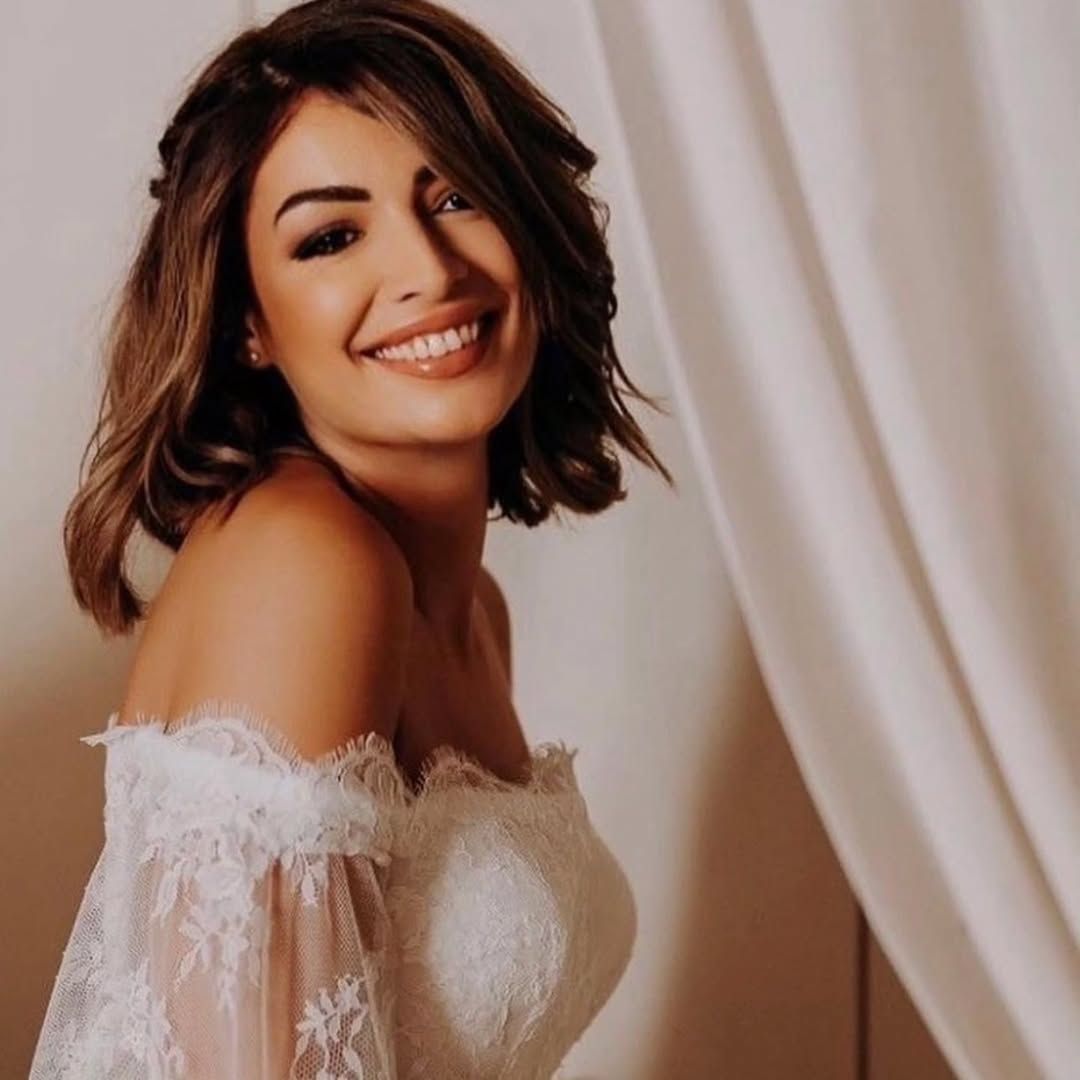 Donna con i capelli castani, sorridente, che indossa un abito di pizzo con le spalle scoperte, accanto a una tenda bianca.