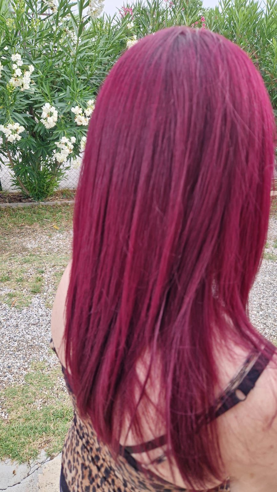 Donna con capelli lunghi, lisci, color magenta, che indossa un top fantasia, all'aperto con fiori bianchi sullo sfondo.