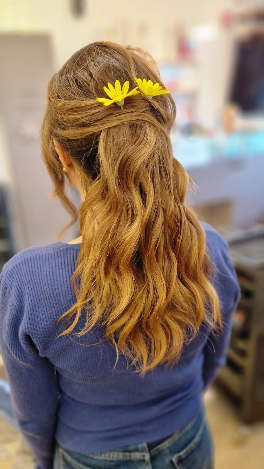 Donna con capelli castani ondulati, raccolti in una mezza coda di cavallo con fermagli a forma di fiore giallo; indossa un maglione blu.