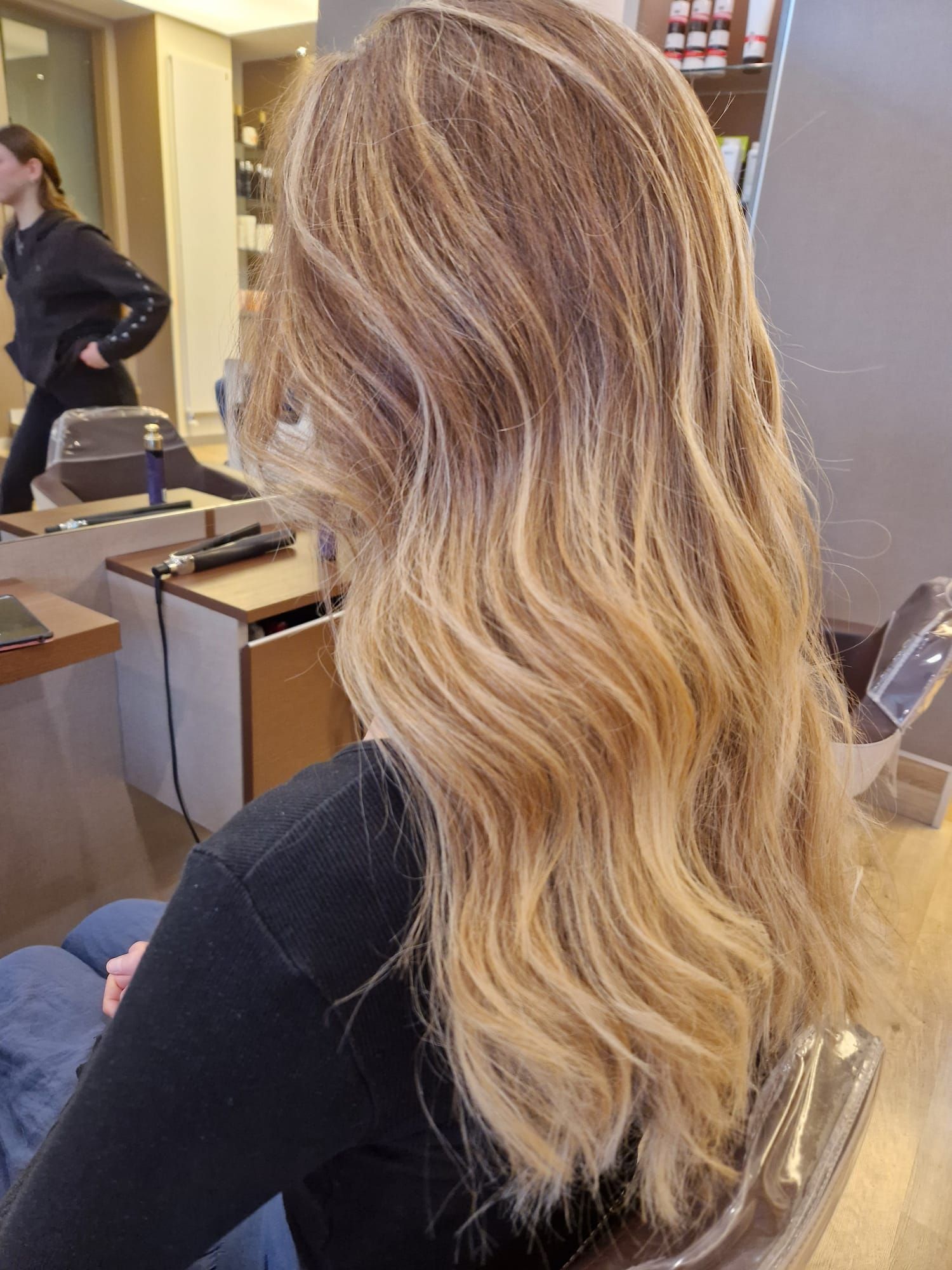 Donna con capelli biondi ondulati, seduta in un salone, di spalle. Capelli con mèches, illuminazione intensa.