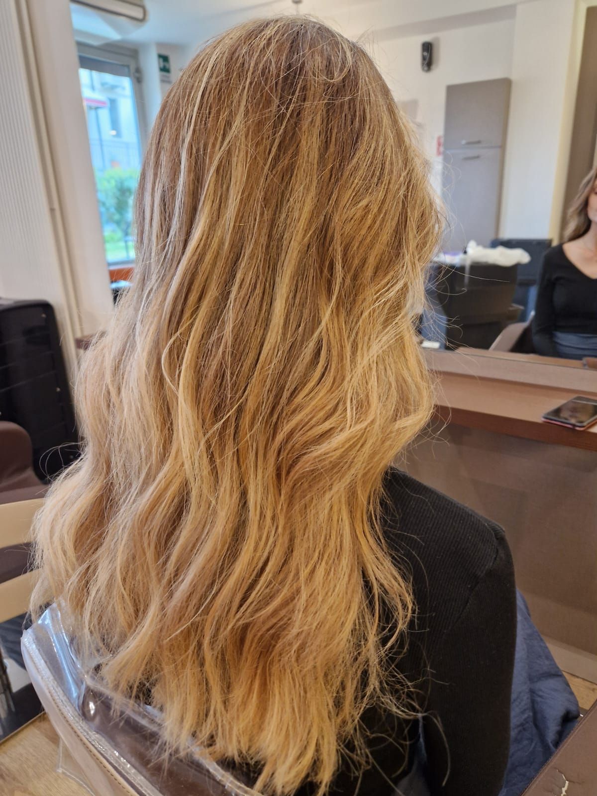 Donna con lunghi capelli biondi e ondulati, acconciata in un salone di bellezza, che dà le spalle alla telecamera.