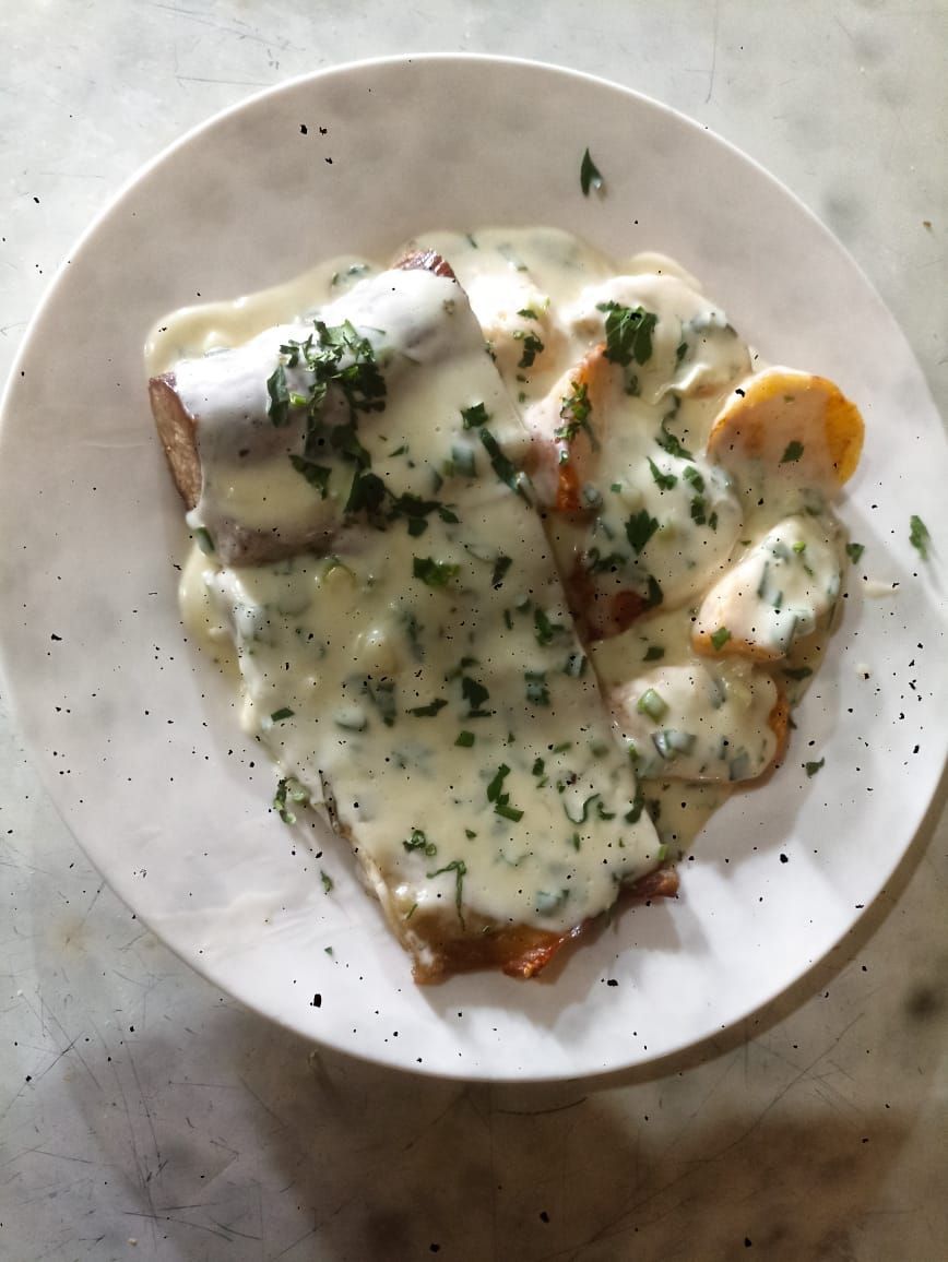 Plato blanco con carne empanizada y rodajas de papa cubiertas con una salsa cremosa con perejil picado.